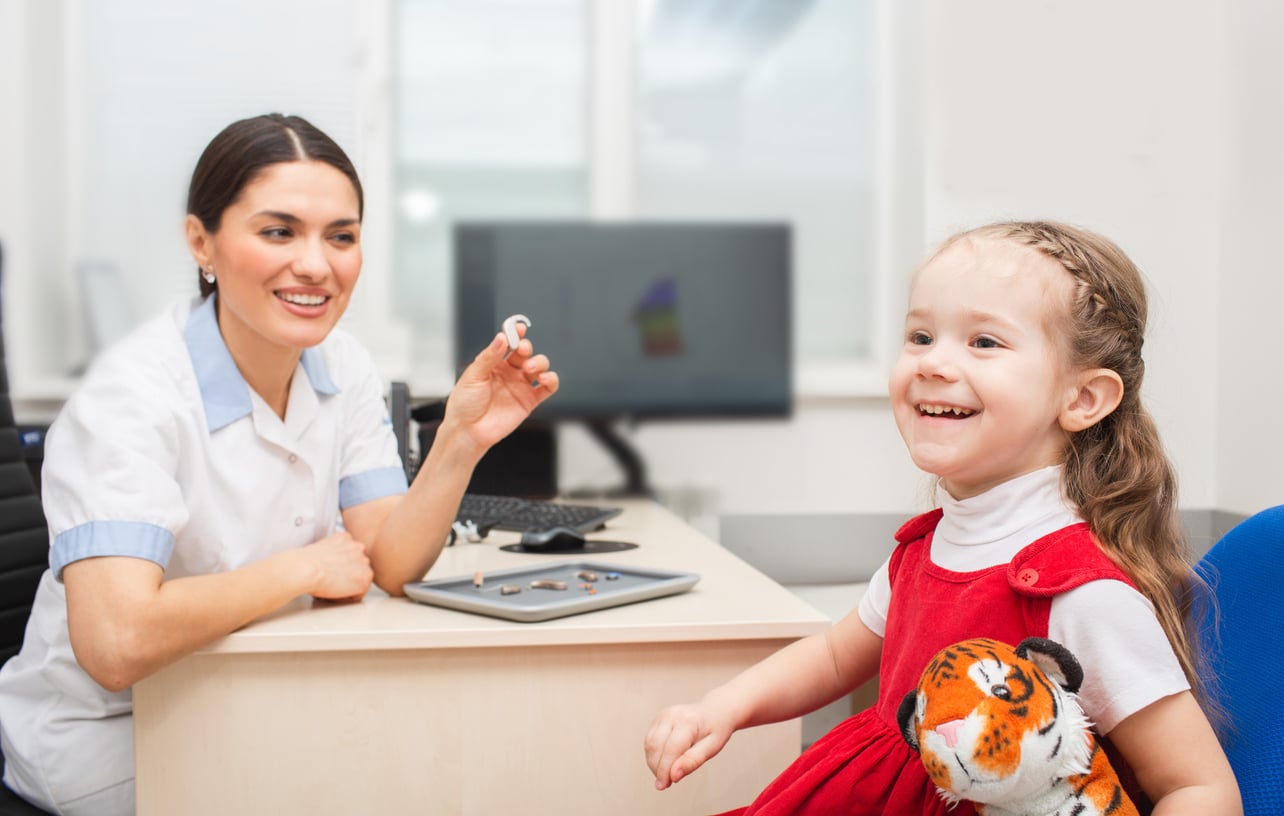 Audiologist explaining hearing aids to smiling young girl.