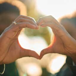 Loving couple expressing affection and unity by forming a heart shape with their hands, backlighting from golden hour sun creating a warm, romantic glow