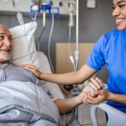 A senior man lies in a hospital bed, smiling and interacting with a dedicated healthcare worker. The room is equipped for medical support and recovery.