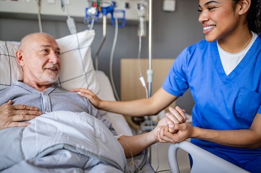 A senior man lies in a hospital bed, smiling and interacting with a dedicated healthcare worker. The room is equipped for medical support and recovery.